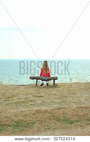 A Little Girl Sits Alone. Solo Travelling. A Tourist Sits On A Wooden Bench On A Cliff Above The Sea