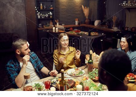 Multi-ethnic Group Of Young People Playing Guessing Game While Sitting At Table During Dinner Party 