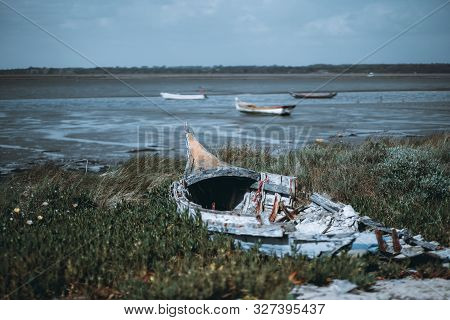 An Old Dilapidated Wooden Hull With Metal Frames Of A Fishing Boat On The Grass Of The River Bank Wi