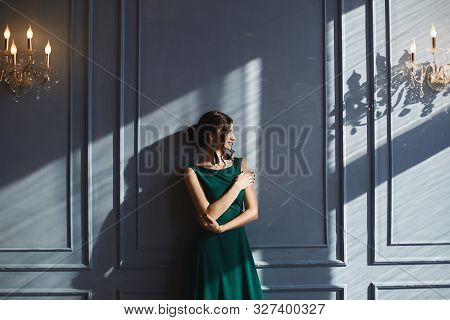 A Beautiful Brunette Girl With Modish Hairstyle, In Green Evening Dress, Stands At The Blue Wall Wit