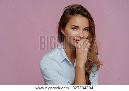 Photo Of Self Assured Attractive Woman With Long Hair, Keeps Hand On Lips, Dressed In Blue Shirt, Lo