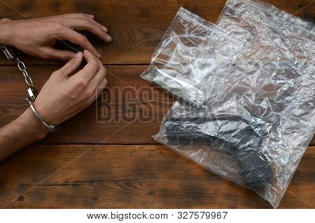 Handcuffed Hands Of Criminal Suspect On Wooden Table And Handgun With Jackknife In Transparent Plast