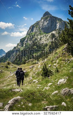 Hikers Walking On Trail. Group Of People Hiking In Mountains With Blooming Meadows And Mountain Peak