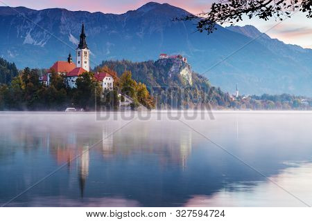 Beautiful Morning At Lake Bled During Sunrise. Mist Lingering Over The Lake With Catholic Church On 