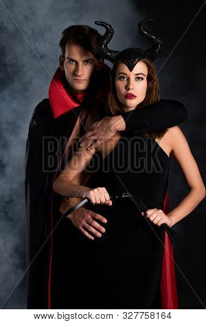 Handsome Man In Cloak Hugging Girl With Horns Holding Flogging Whip On Black With Smoke