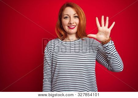 Young redhead woman wearing strapes navy shirt standing over red isolated background showing and pointing up with fingers number five while smiling confident and happy.