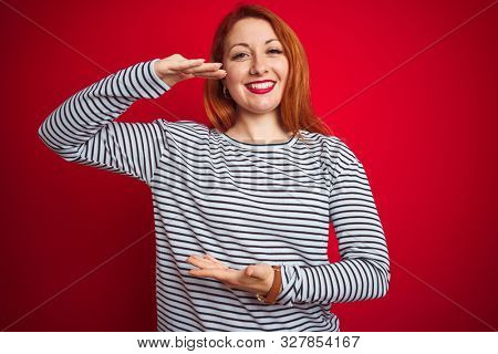 Young redhead woman wearing strapes navy shirt standing over red isolated background gesturing with hands showing big and large size sign, measure symbol. Smiling looking at the camera.