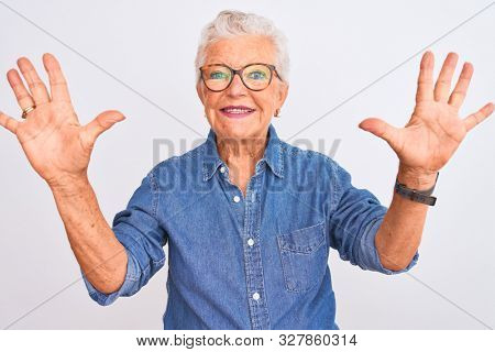 Senior grey-haired woman wearing denim shirt and glasses over isolated white background showing and pointing up with fingers number ten while smiling confident and happy.