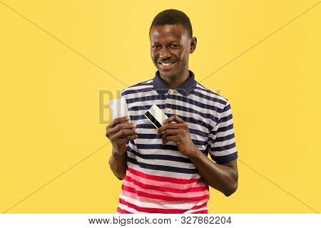 Young African-american Man With Smartphone And Debet Card Isolated On Yellow Studio Background, Faci