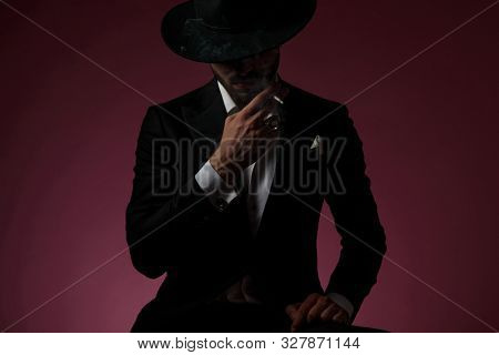 Powerful young man smoking a cigarette and leaning on his leg while wearing a black suit and hat, sitting on cherry-colored studio background