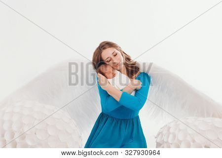 A Girl In A Blue Dress With Angel Wings Holding A Newborn Baby On A White Background. An Angel-woman