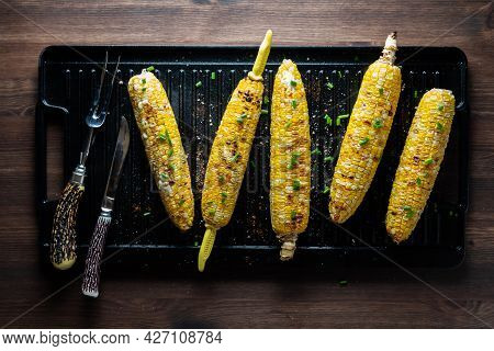 Top Down View Of Corn On The Cob On A Black Grill Pan, Topped With Seasoning And Herbs.