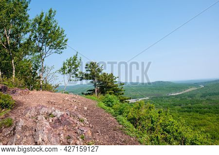 Summer Landscape, Pine Trees On A Rock Around A Green Forest And A Road In The Distance