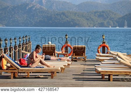 Hisaronu, Turkey - July 2021: Smiling Girl In Bikini Sitting With Smartphone In Sunbed On A Beach On
