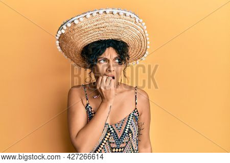 Young hispanic woman holding mexican hat looking stressed and nervous with hands on mouth biting nails. anxiety problem. 