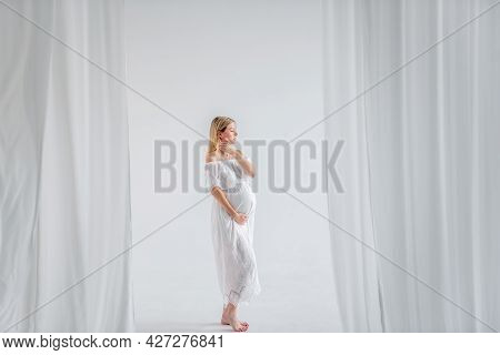 A Young Pregnant Woman In Anticipation Of A Baby In A White Dress, Hugs Belly. Fashion Girl In A Min