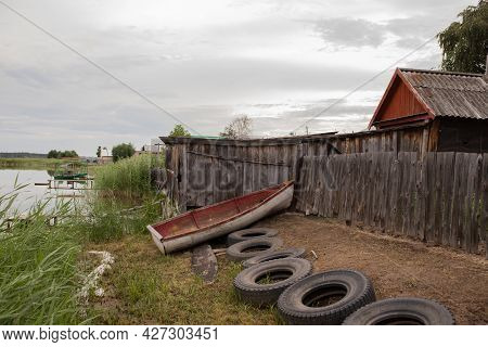 An Old Wooden Boat Sits On The Shore Next To An Old House On The Lake. Old Wooden House On The Shore