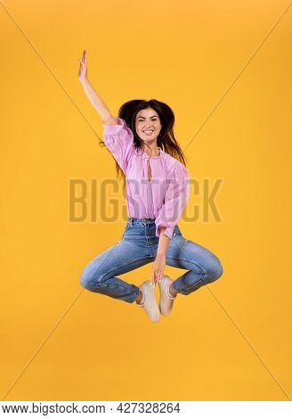 Joyful Armenian Woman Jumping And Touching Legs With Hand, Yellow Studio Background, Full Length Pho