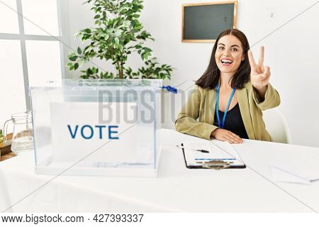 Young brunette woman sitting at election table with voting ballot showing and pointing up with fingers number two while smiling confident and happy. 