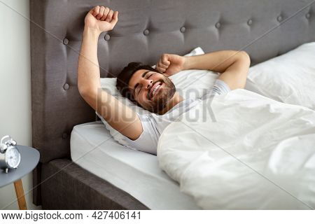 Rested Arab Man Waking Up And Stretching Hands, Wellslept Guy Lying In Bed, Awakening In Cozy Bedroo