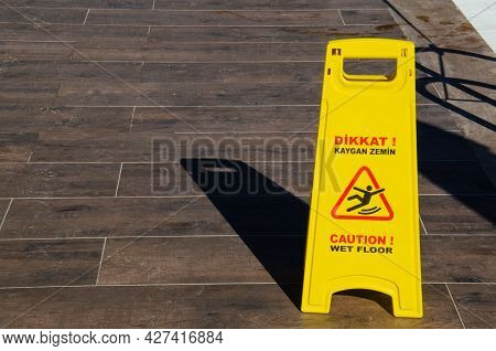Yellow Warning Sign For Wet Floor Standing On Brown Tiled Floor. The Inscription On Yellow Informati