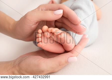 Female Hands Of Young Mother Holding Her Newborn Baby Feet, Closeup Image With Blur Baby In Backgrou