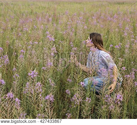 Beautiful Smiling Blond Young Woman In Light Purple Shirt Sitting On Field Of Fireweed Flowers Sniff