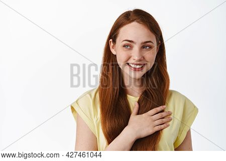 Tender And Cute Redhead Female Model Holding Hand On Heart, Looking Aside At Logo With Heartwarming 