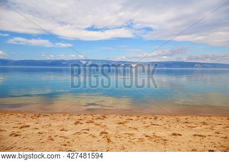 Beautiful Summer Background. Sandy Beach, Blue Water, Mountains In The Background, Cloudy Sky. Lake 