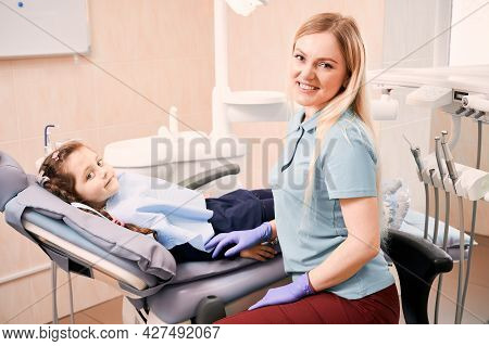 Adorable Kid Looking At Camera And Smiling While Lying In Dental Chair. Woman Dentist In Sterile Glo