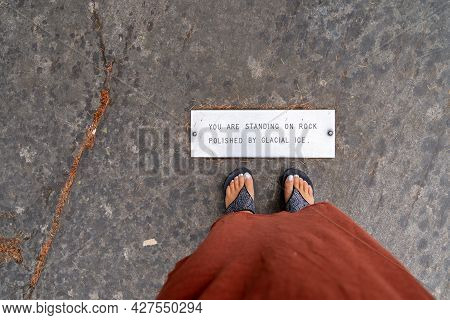 Woman Tourist Wearing Rhinestone Flip Flop Shoes Stands Near A Sign, Standing On Rock Polished By Gl