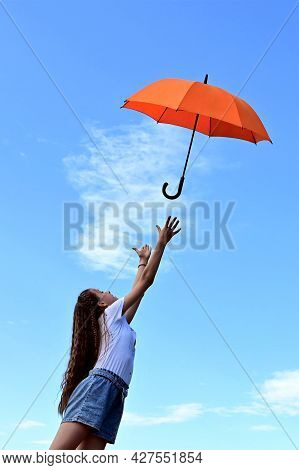 Beautiful Young Girl In A White T-shirt And Denim Shorts Throws Up An Orange Umbrella. A Flying Umbr