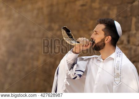 Jewish Man Blowing Shofar On Rosh Hashanah Outdoors. Wearing Tallit With Words Blessed Are You, Lord