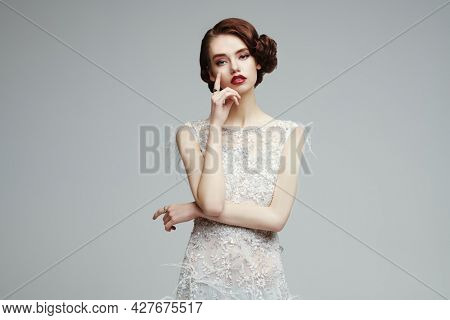 Evening makeup and hairstyle. Portrait of a charming young woman in a luxury white dress on a gray background. Hair and makeup in the style of the 20s. 