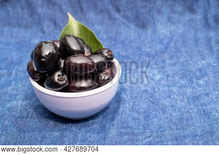 Organic Jamun Or Black Plum In A Bowl Isolated On Blue Fabric Background With Leaf, Also Known As Ma