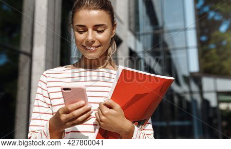 Portrait Of Smiling Blond Girl Chatting On The Way To Universy Or Work. Woman Holding Books And Look