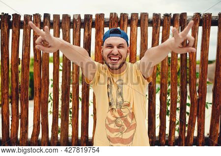 Cheerful Bearded Young Boy In Yellow T Shirt Showing Peace Gesture With Both Hands, Smiling And Lean