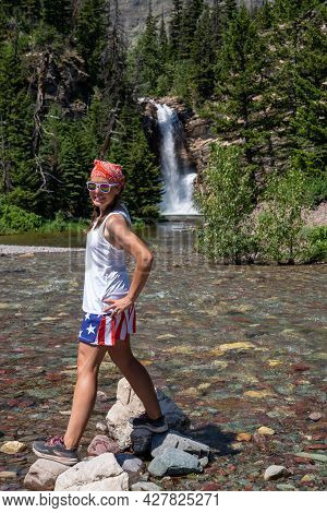 Young Adult Woman Wearing American Flag Patriotic Clothing Hikes And Crosses The Rocks And Stream By