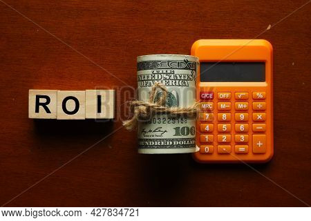 Roi Wooden Block With Fake Money And Calculator. Roi Stands For Return Of Investment.