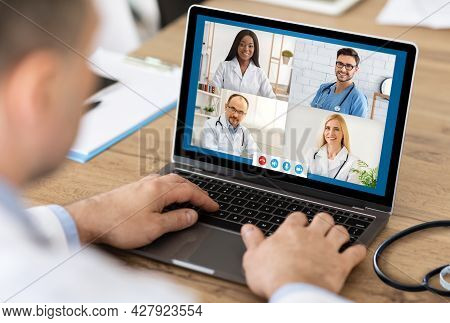 Senior Male Doctor Consulting Colleagues Via Video Call At Workplace