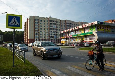 Koltsovo, Novosibirsk, Russia - 24 July, 2021: Car Cadillac stops in front of pedestrian crossing for woman with bicycle crosses road near department store, Work settlement of Koltsovo, Roads Siberia