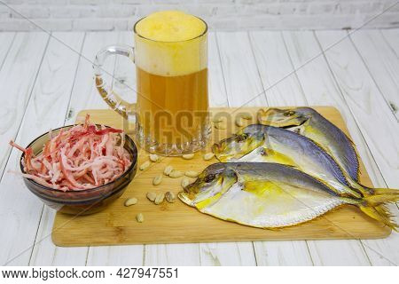 A Mug Of Beer With Foam, Dried Vomer Fish And Squid Shavings In A Round Black Plate On A Wooden Boar