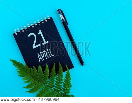 April 21th. Day 21 Of Month, Calendar Date. Black Notepad Sheet, Pen, Fern Twig, On A Blue Backgroun