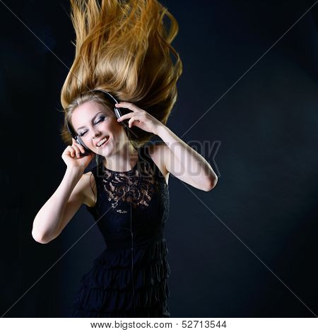 Music girl. Young beautiful excited woman with headphones listening music and dancing with long blond hair fly-away against dark background 