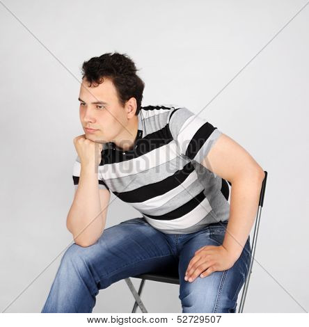 Brunet Man In Striped T-shirt Sits On Chair And Thinks On Grey Background.