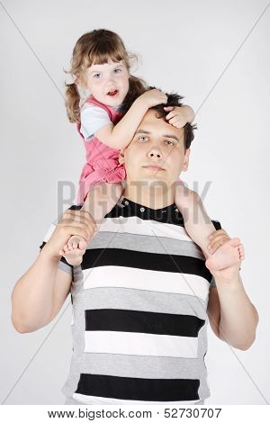 Little Girl Sits On Shoulders Of Her Father And Holds His Hair On Grey Background.