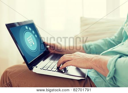 technology, leisure, advertisement and lifestyle concept - close up of man working with laptop computer displaying shopping trolley icon on screen and sitting on sofa at home