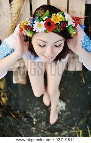 Girl In A Flower Wreath On His Head Sitting On The Bridge And Wets Feet In The River