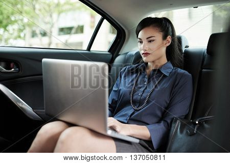 Pretty young business lady sitting on backseat of car and working