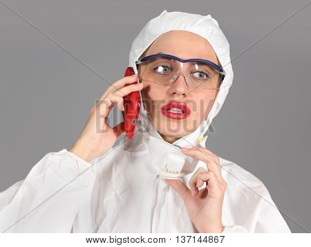 headshot of woman in white protective suit of synthetic paper, glasses and respiritory half mask, talking on phone, disquieted, on gray background
